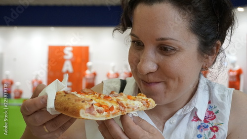 Girl eating pizza. Sitting in the food zone at the Mall. On the background of the store. Light bright clothes. A hungry greedy look. Close-up portrait.