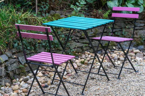 Two pink wooden folding chairs and a square blue table are standing on gravel in the backyard of a cafe in natural light in the evening