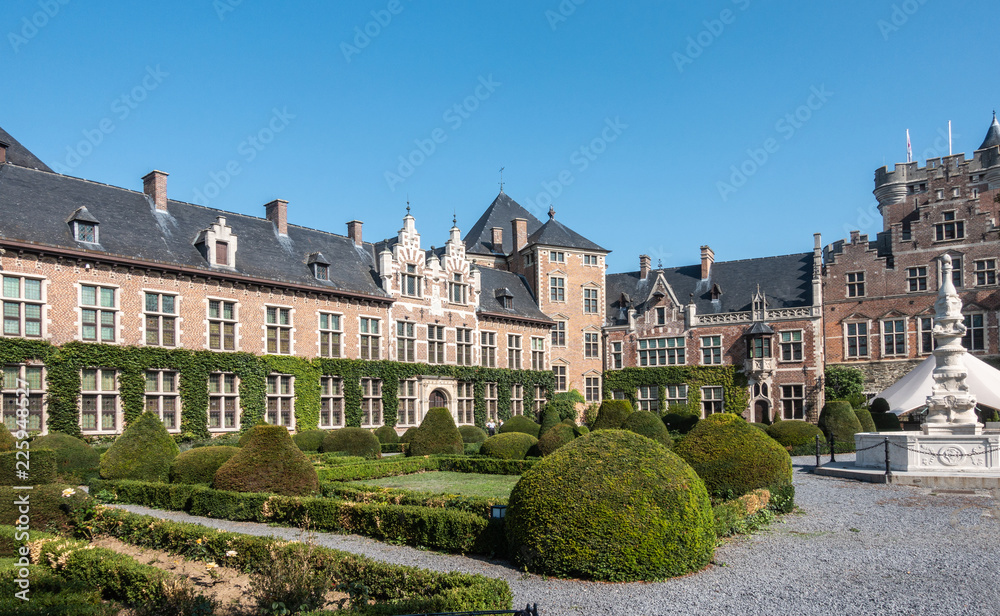 Gaasbeek, Flanders, Belgium - September 14, 2018: Southwest wing and entrance gate and tower of Gaasbeek Castle with green garden in front, light brown walls, dark roof under blue sky.