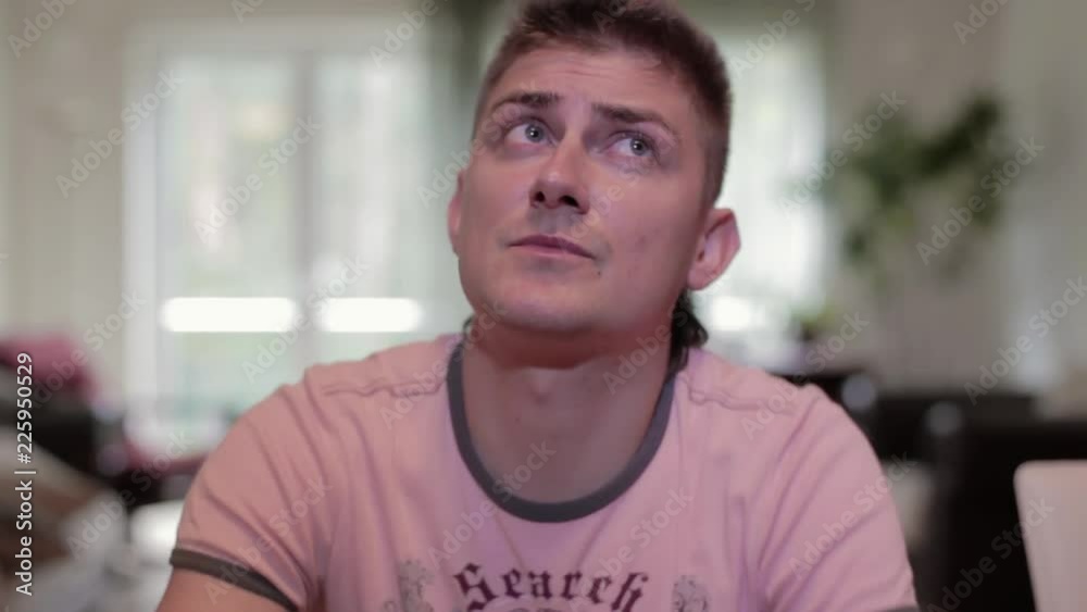 Young caucasian handsome grey-eyed guy with modern haircut dressed in pink t-shirt stares up at ceiling with worried face, on blurry background of windows with white curtains. Young caucasian handsome grey-eyed guy with modern haircut dressed in pink t-shirt stares up at ceiling with worried face, on blurry background of windows with white curtains.