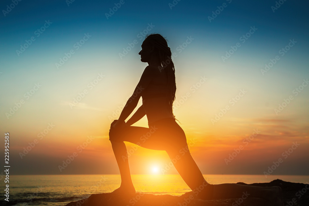 Silhouette of a young woman practicing yoga exercises on the ocean at amazing sunset.