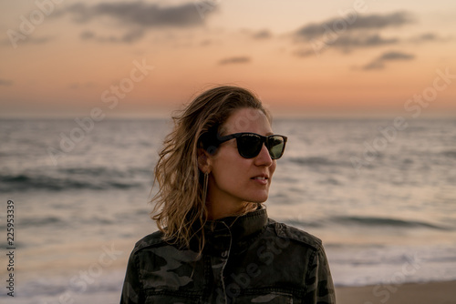 Beautiful blond curly hair woman wearing camouflaged jacket and sunglasses posing at sunset on the beach © Henrique