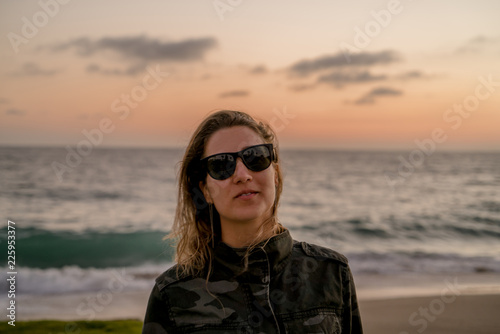 Beautiful blond curly hair woman wearing camouflaged jacket and sunglasses posing at sunset on the beach © Henrique