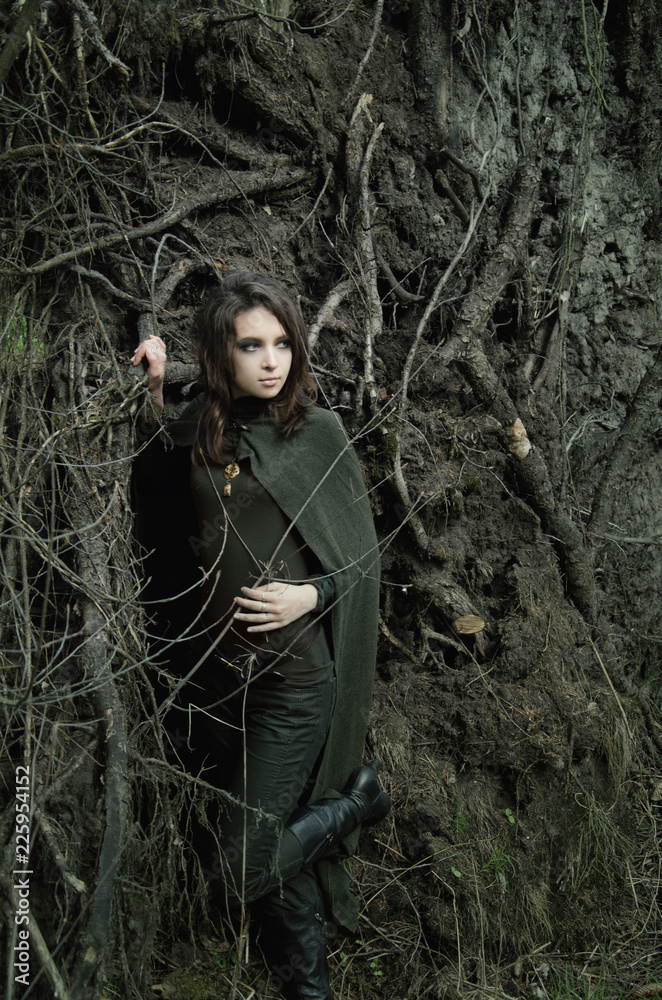 portrait of a young woman in an elven cloak standing among the roots of trees in a dark forest