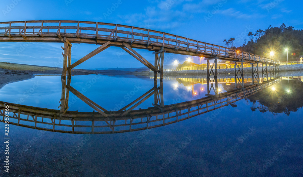Obraz premium Amazing landscape at twilight in Buchupureo, a below view of the bridge that cross the river to access the beach and its reflection on the water illuminated by the blue hour and the first lamps, Chile