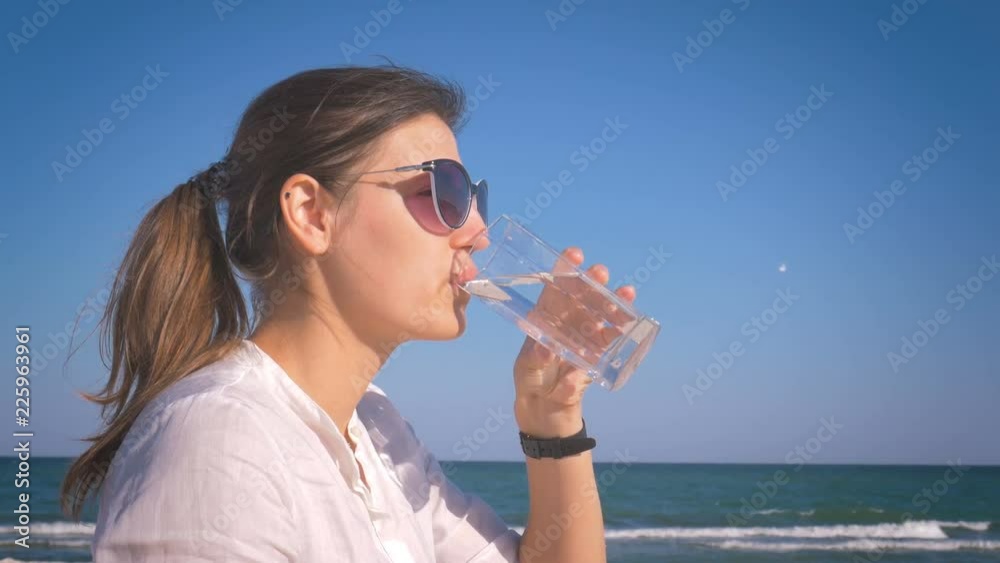 Girl in a white shirt and sunglasses sipping water from a glass and smiling sitting on the sea shore Girl in a white shirt and sunglasses sipping water from a glass and smiling sitting on the sea shore