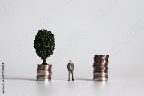 Photography Miniature man standing between a pile of coins and a tree on a pile of coins