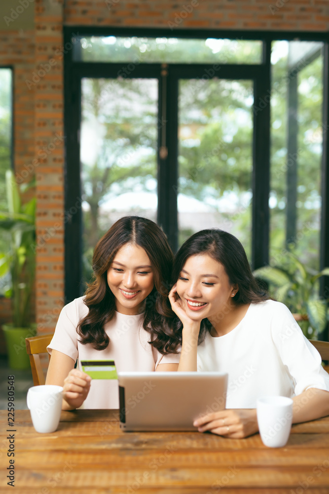 Happy relaxed young female friends doing online shopping through laptop and credit card in living room Happy relaxed young female friends doing online shopping through laptop and credit card in living room
