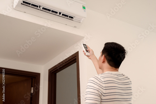 Young man switching on or adjusting the wall mounted air conditioner in the living room with a remote control photo
