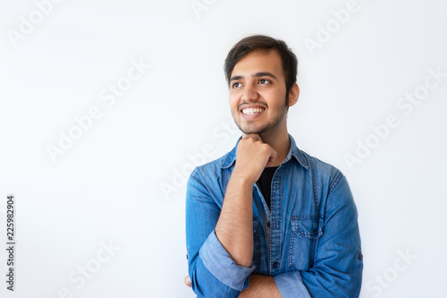 Portrait of young Indian man looking upwards and smiling. Positive handsome dark haired man wearing denim shirt propping up chin with right hand. Happy memory concept.