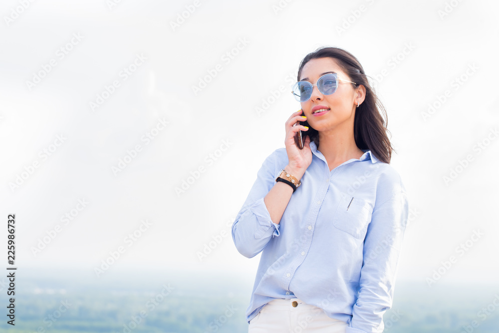 Happy fashionable woman with glasses talking on the phone and smiling at the background of nature, sky. Business lady is successful in negotiations Happy fashionable woman with glasses talking on the phone and smiling at the background of nature, sky. Business lady is successful in negotiations