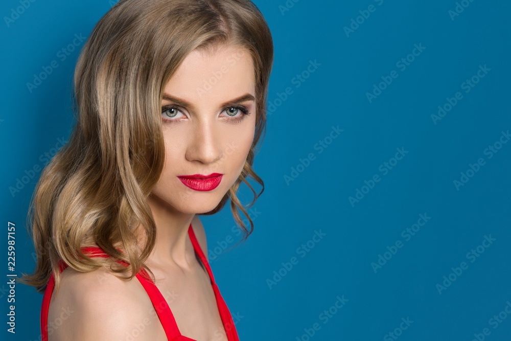 Beauty portrait of young blonde woman with bright red lips, blue eyes, in red dress on blue background, copy space Beauty portrait of young blonde woman with bright red lips, blue eyes, in red dress on blue background, copy space