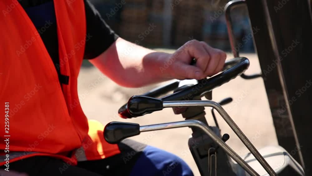 The worker on the fork loader. The worker operates the fork loader. The wheel, control levers are largely removed. The employee is visible partially. On him overalls. There is no face of the employee The worker on the fork loader. The worker operates the fork loader. The wheel, control levers are largely removed. The employee is visible partially. On him overalls. There is no face of the employee
