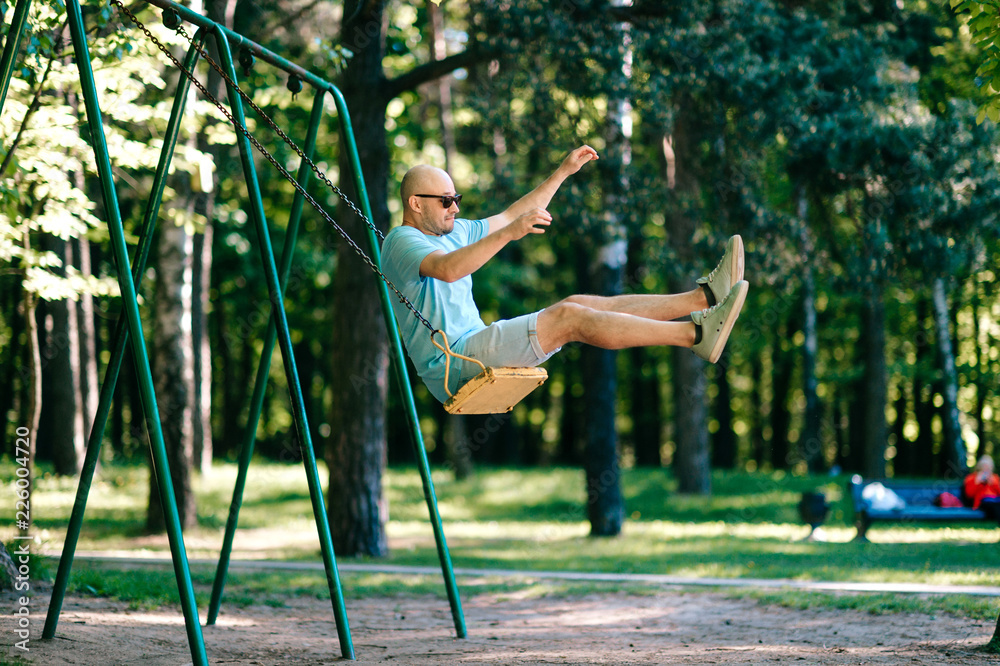 Odd adult bald man in sunglasses jumping from swing on ground in motion.  Childhood behavior. Hulking awkward foolish comic male have fun on children playground outdoor in city park in summer day.