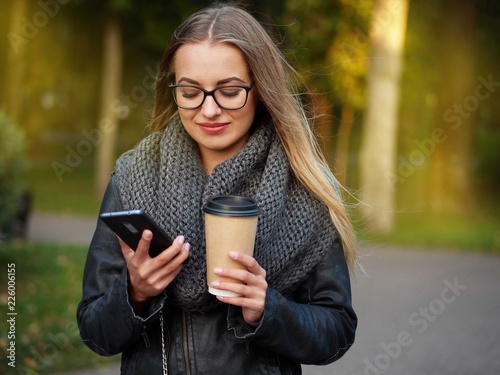 Portrait of a beautiful stylish young blonde with make-up in black leather jacket glasses and a knitted scarf. Frozen girl drinks hot drink and talks on the phone in the autumn cold park