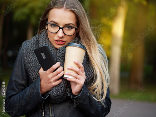 Portrait of a beautiful stylish young blonde with make-up in black leather jacket glasses and a knitted scarf. Frozen girl drinks hot drink and talks on the phone in the autumn cold park