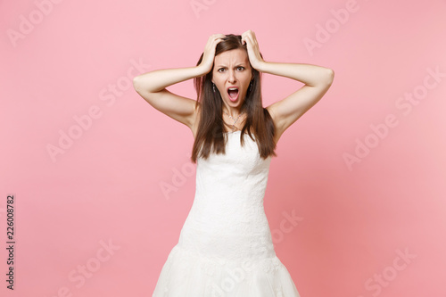 Portrait of irritated dissatisfied bride woman in white wedding dress standing screaming clinging to head isolated on pink pastel background. Wedding celebration concept. Copy space for advertisement.
