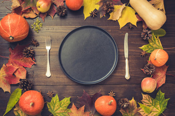 Autumn and Thanksgiving day table setting with fallen leaves, pumpkins, spices, empty black platter and vintage cutlery on brown wooden table. Top view, toned image.