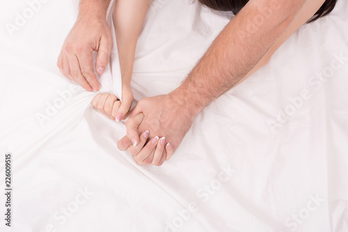 Hands of couple who making love in bed on white crumpled sheet, focus on hands