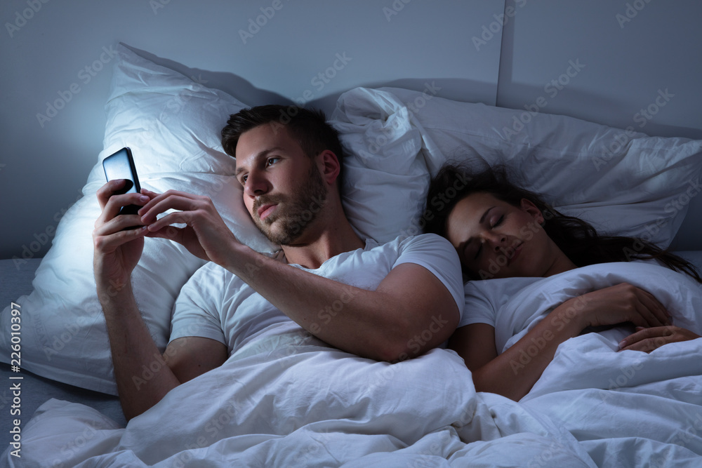 Man Using Cellphone While Her Wife Sitting On Bed - obrazy, fototapety, plakaty 