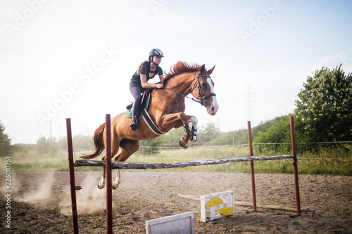 Slika na platnu Young female jockey on horse leaping over hurdle