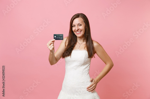 Portrait of smiling bride woman in beautiful lace white wedding dress holding credit card isolated on pink pastel background. Organization of wedding celebration concept. Copy space for advertisement.