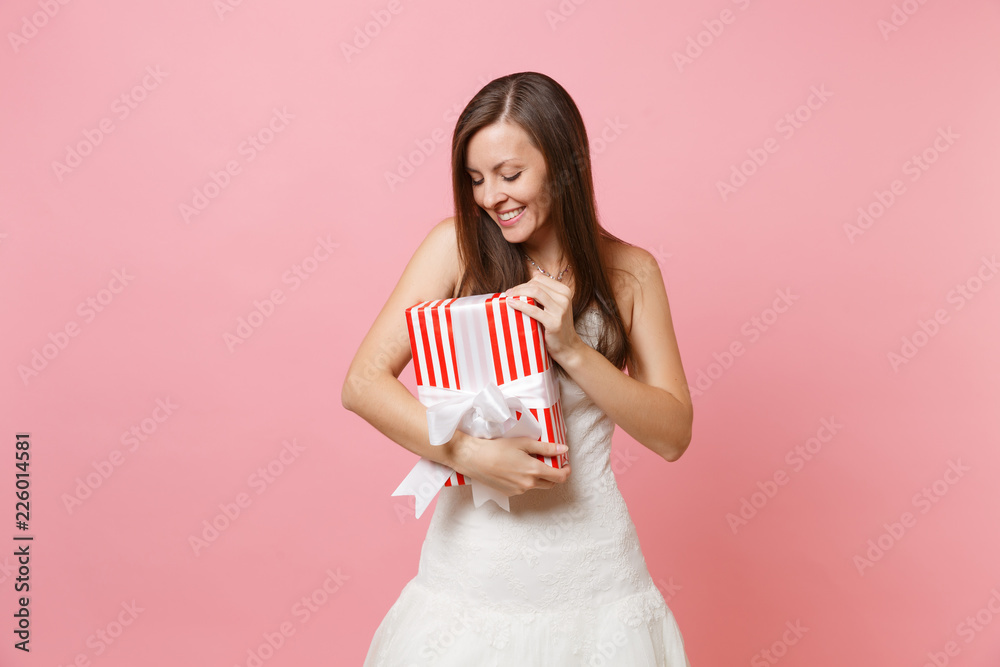 Portrait of tender bride woman in beautiful lace white wedding dress looking on red box with gift present isolated on pink pastel background. Wedding celebration concept. Copy space for advertisement. Portrait of tender bride woman in beautiful lace white wedding dress looking on red box with gift present isolated on pink pastel background. Wedding celebration concept. Copy space for advertisement.