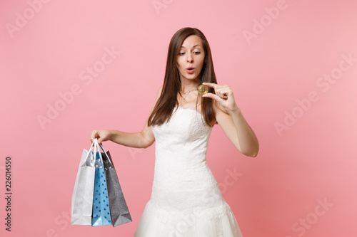 Surprised bride woman in wedding dress looking on bitcoin metal coin of golden color hold multi colored packages bag with purchases after shopping isolated on pink background. Organization of wedding.