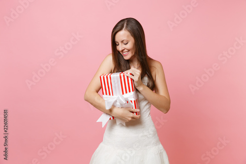 Portrait of tender bride woman in beautiful lace white wedding dress looking on red box with gift present isolated on pink pastel background. Wedding celebration concept. Copy space for advertisement.