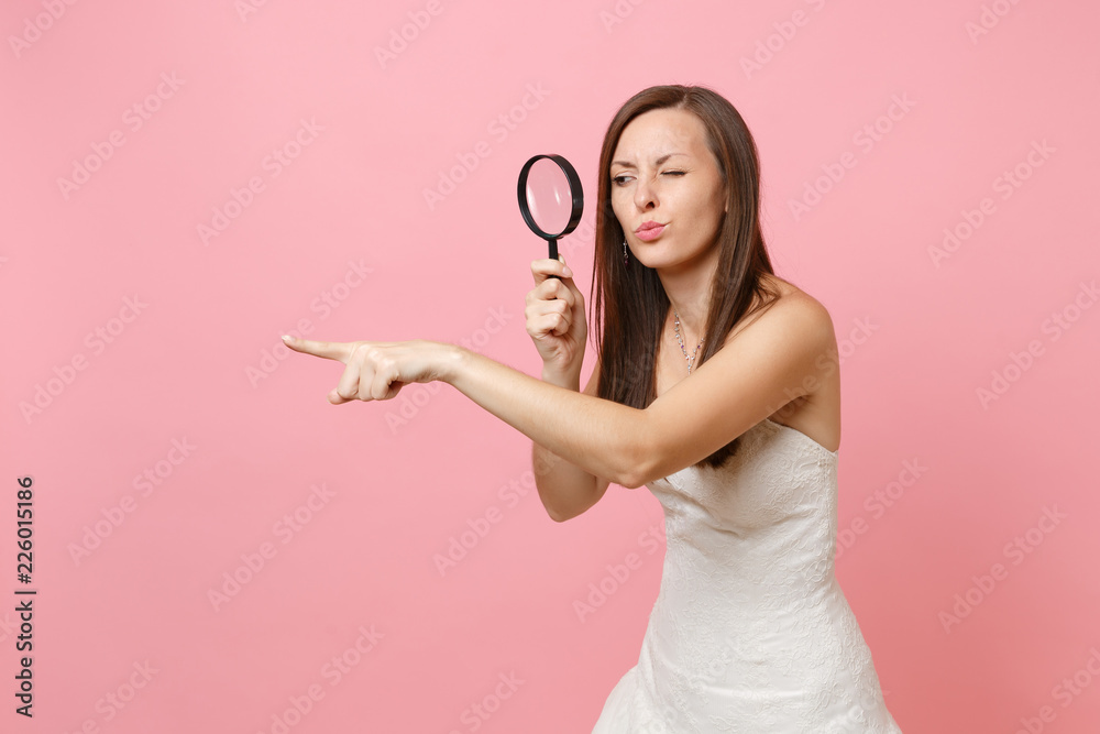 Concerned dissatisfied bride woman in wedding dress pointing index finger aside, looking through magnifying glass isolated on pastel pink background. Wedding to do list. Organization of celebration. Concerned dissatisfied bride woman in wedding dress pointing index finger aside, looking through magnifying glass isolated on pastel pink background. Wedding to do list. Organization of celebration.