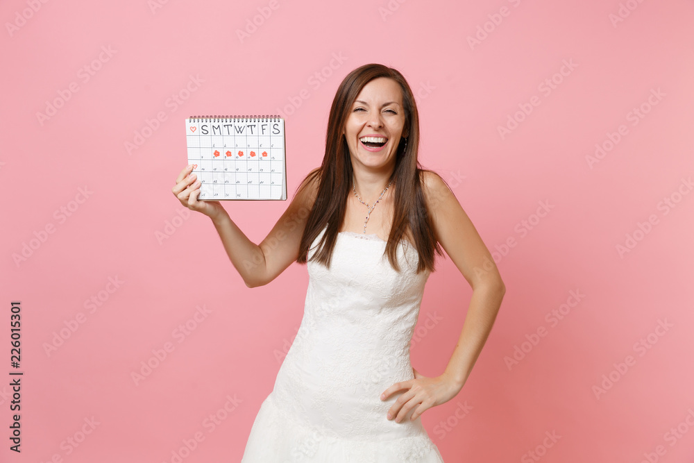 Laughing bride woman in white wedding dress holding female periods calendar for checking menstruation days isolated on pastel pink background. Medical, healthcare, gynecological concept. Copy space. Laughing bride woman in white wedding dress holding female periods calendar for checking menstruation days isolated on pastel pink background. Medical, healthcare, gynecological concept. Copy space.