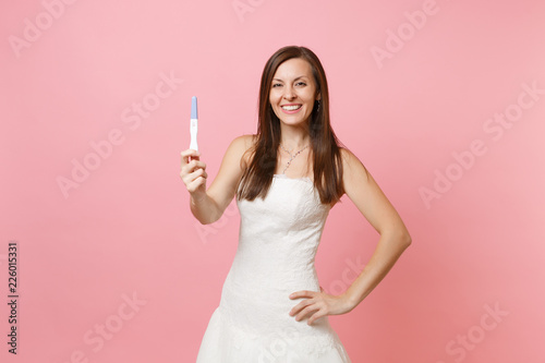 Smiling joyful bride woman in white wedding dress holding pregnancy test isolated on pastel pink background. Medical healthcare gynecological pregnancy fertility maternity people concept. Copy space.