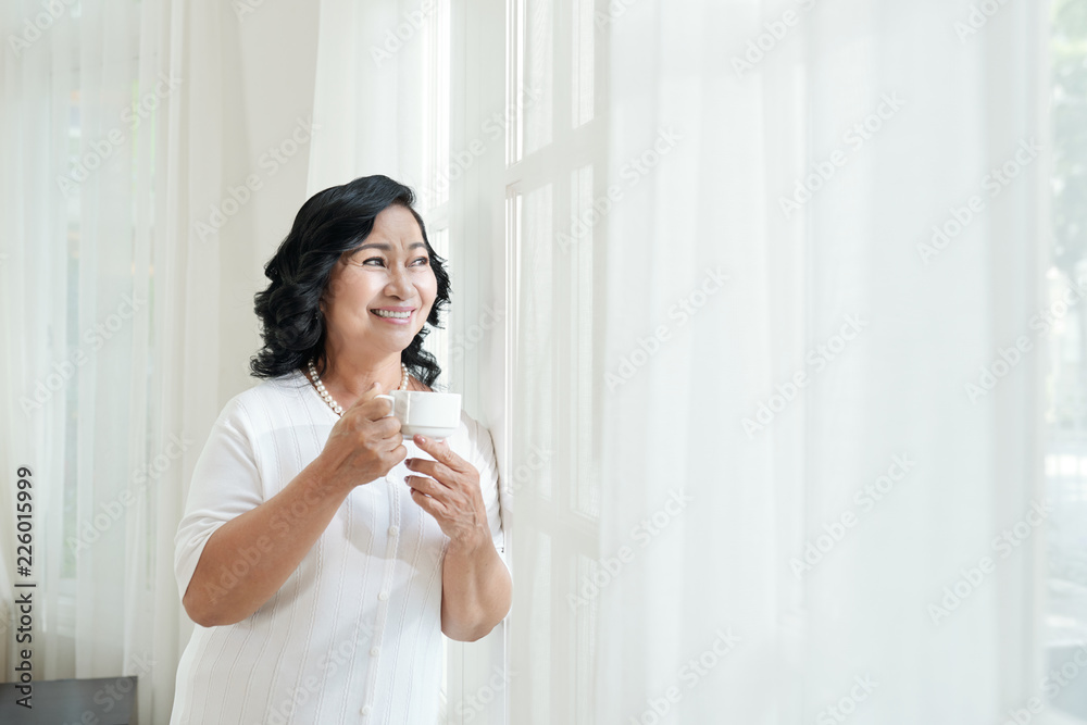 Portrait of beautiful elderly Asian woman holding cup of fresh coffee and smiling cheerfully while looking out the window Portrait of beautiful elderly Asian woman holding cup of fresh coffee and smiling cheerfully while looking out the window