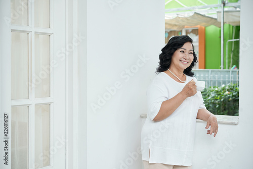 Portrait of elegant beautiful mature Asian woman standing on porch with cup of tea, looking away and smiling happily