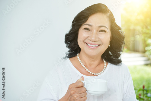 Waist-up portrait of beautiful senior Asian woman with pearl necklace holding cup of tea and smiling at camera cheerfully