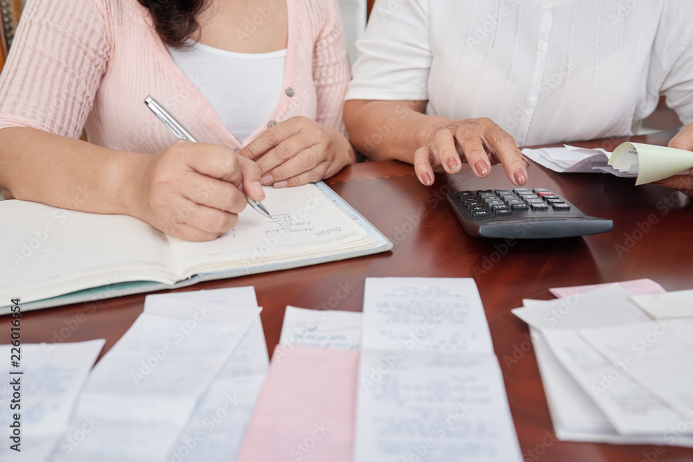 Crop shot of unrecognizable mature women sitting at table with receipts, using calculator and writing down notes in notepad Crop shot of unrecognizable mature women sitting at table with receipts, using calculator and writing down notes in notepad