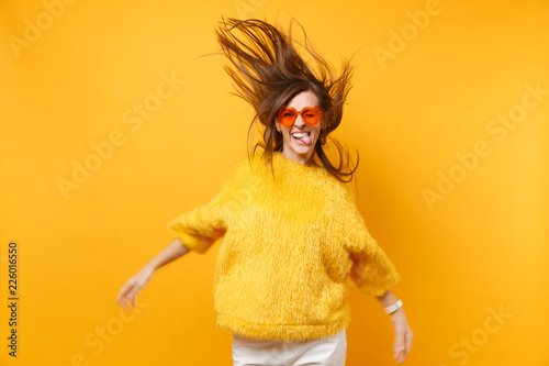Comic young girl in fur sweater heart orange glasses showing tongue, fooling around in studio jump with flying hair isolated on yellow background. People sincere emotions, lifestyle. Advertising area.