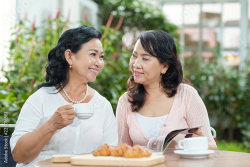 Beautiful elegant mature Asian women sitting at cafe table outdoors, drinking coffee with croissants and enjoying friendly talk