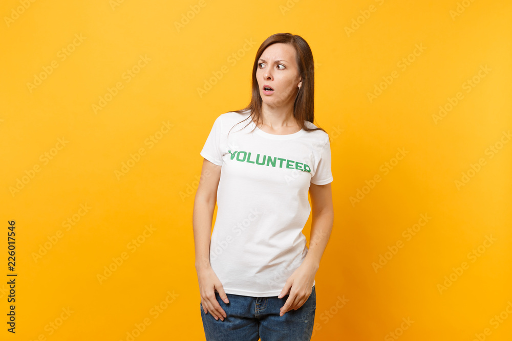 Portrait of scared shocked afraid young woman in white t-shirt with written inscription green title volunteer isolated on yellow background. Voluntary free assistance help, charity grace work concept. Portrait of scared shocked afraid young woman in white t-shirt with written inscription green title volunteer isolated on yellow background. Voluntary free assistance help, charity grace work concept.