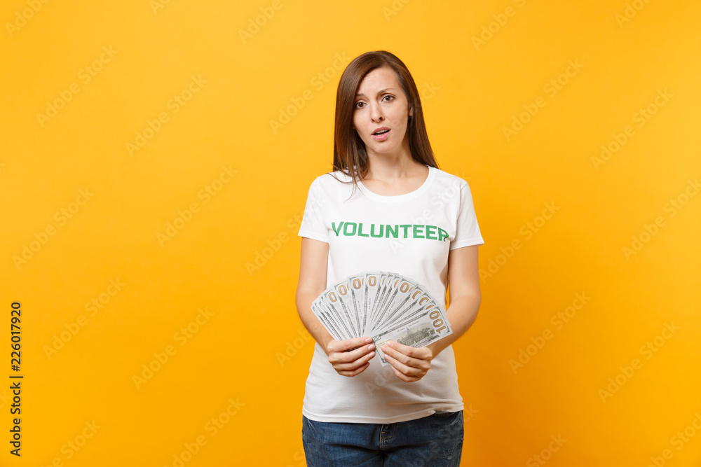Woman in white t-shirt with written inscription green title volunteer hold lots dollars banknotes, cash money isolated on yellow background. Voluntary free assistance help, charity grace work concept. Woman in white t-shirt with written inscription green title volunteer hold lots dollars banknotes, cash money isolated on yellow background. Voluntary free assistance help, charity grace work concept.