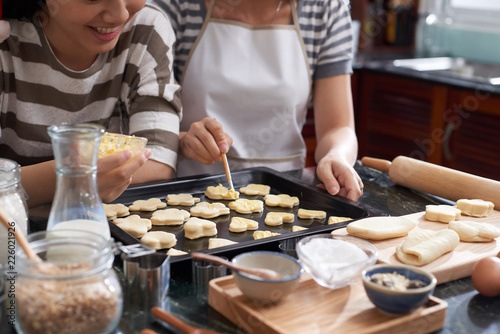 Crop shot of unrecognizable women standing by kitchen table and spreading butter on cut out raw cookies lying on baking sheet