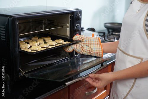 Unrecognizable woman wearing oven mitt putting baking sheet with raw cookies into modern oven in home kitchen