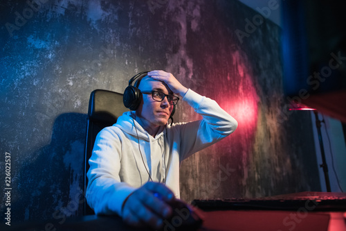 Angry young gamer in glasses playing game on computer using headphones photo