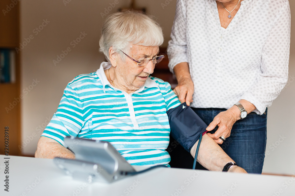 Senior woman measuring blood pressure at home with help of other woman Senior woman measuring blood pressure at home with help of other woman