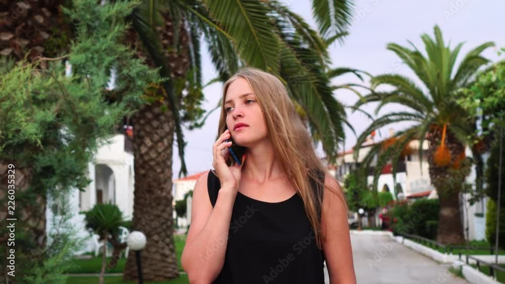 Young beautiful blonde female smiling while walking slowly talking on smartphone at tropical palm tree resort vacation Young beautiful blonde female smiling while walking slowly talking on smartphone at tropical palm tree resort vacation