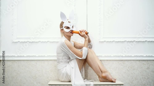 girl in white dress and in white mask of rabbit eat carrot on white background. Fashionable girl in a hare mask. Sexy Bunny Girl.
