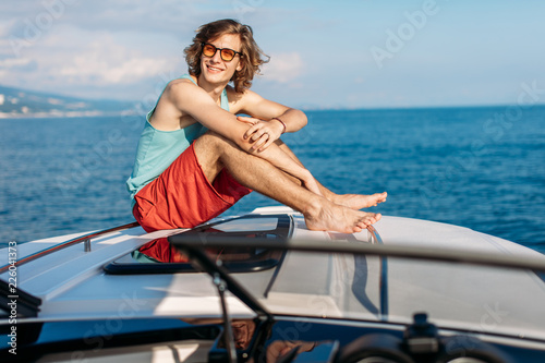 Young caucasian young man on vacation sailing in the sea, sitting on a bow of a motor boat , looking at camera