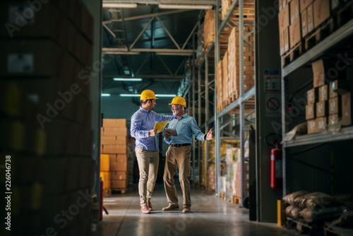 Picture of two business man standing in warehouse with helmets on their heads and celebrating great news about their business. Standing in big warehouse and looking happy and satisfied.