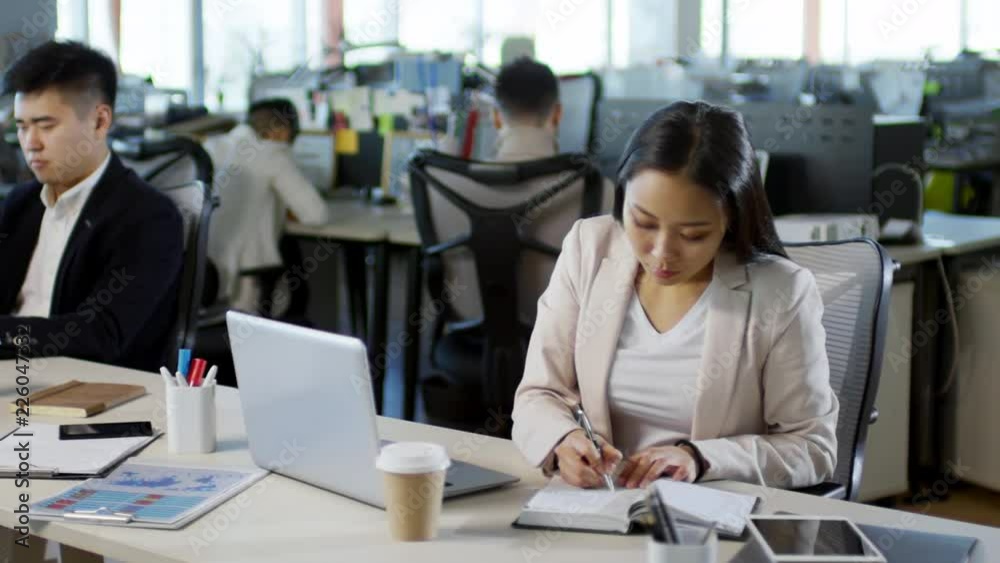 Tracking shot of busy Asian office worker sitting at desk, drinking coffee and writing in notebook when using laptop computer, other people working in the background Tracking shot of busy Asian office worker sitting at desk, drinking coffee and writing in notebook when using laptop computer, other people working in the background