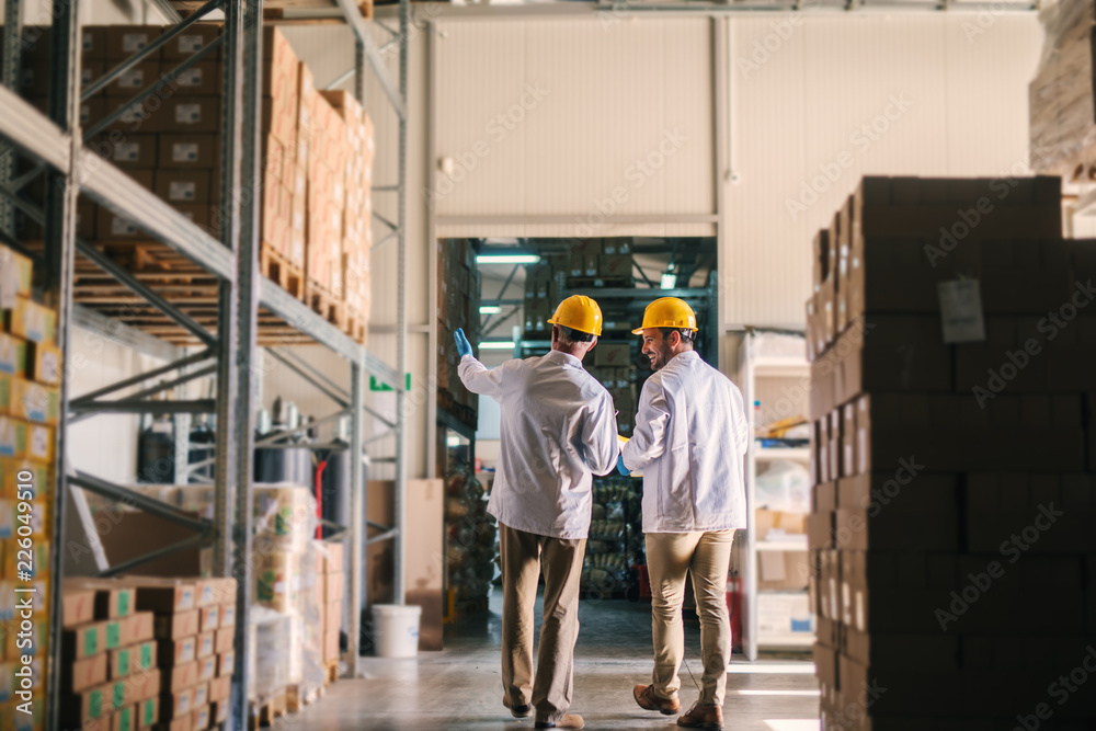 Father and son standing in their warehouse with helmets on their heads and looking at package prepared for transport. Looking proud and satisfied.